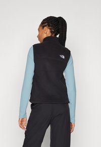 Black fleece vest with a high collar, featuring a small white logo on the back. Styled over a light blue long-sleeve top.