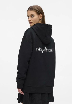 Vrouw in een zwarte hoodie met de tekst "KARL LAGERFELD" en kattenillustraties op de rug, kijkt over haar schouder tegen een witte achtergrond.