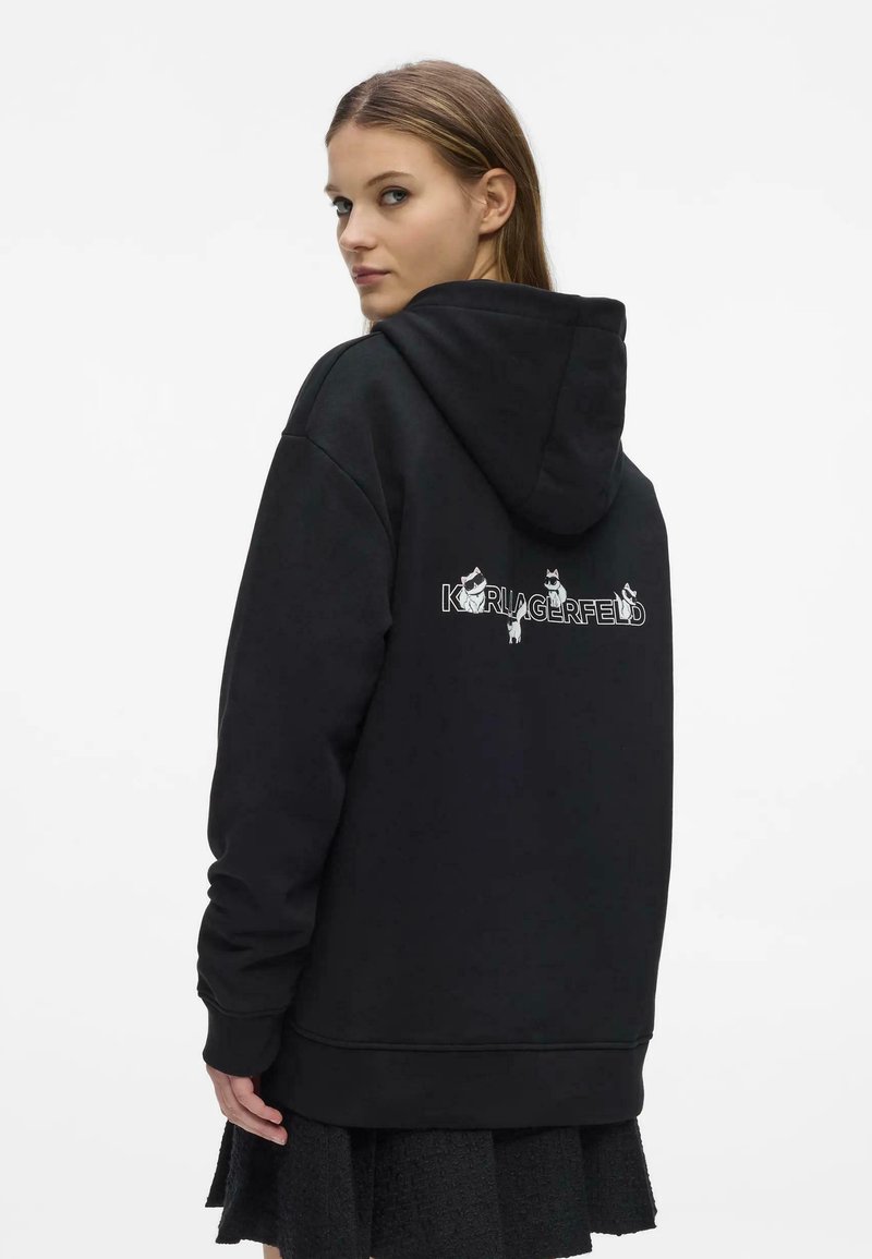 Femme en sweat à capuche noir avec le texte « KARL LAGERFELD » et des illustrations de chats dans le dos, regardant par-dessus son épaule sur fond blanc.