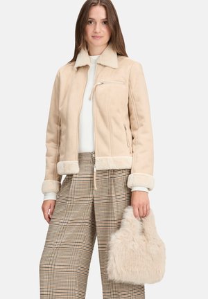 Femme portant une veste en peau lainée beige et un pantalon large à carreaux, tenant un sac à main beige duveteux sur un fond uni.