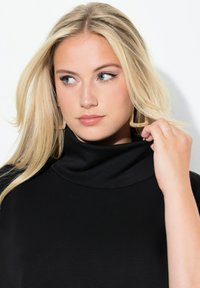 Maglione nero a collo alto in tessuto strutturato; modello con orecchini geometrici dorati, capelli biondo chiaro che incorniciano delicatamente il viso.