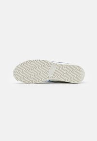 Vitt gummisul av en sneakers med ett zigzag-mönster och rektangulärt logotyppr präglat i centrum på en enfärgad vit bakgrund.