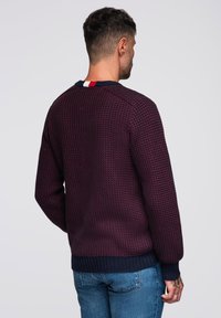 Villa neule tumma purppura, jossa on teksturoitu kuvio. Ominaisuuksiin kuuluu pyöreä pääntie ja kontrastivärinen tummansininen ribbihelma. Värilohkoilla varustettu kaulus.