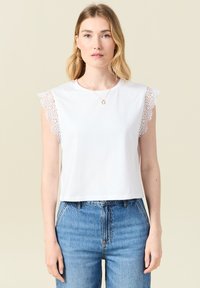 Valkoinen puuvillapusero, jossa on pitsikoristeiset olkapäät ja pyöreä pääntie, yhdistettynä korkeavyötäröisiin sinisiin farkkuihin.