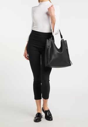 Czarna skórzana torba typu tote o gładkiej fakturze, szerokim otwarciu i bocznych frędzlach, trzymana przez osobę w białej bluzce i czarnych dopasowanych spodniach.