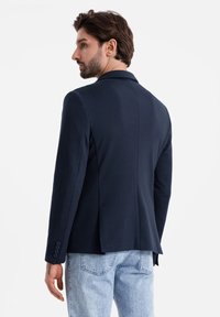 Navy Blazer mit taillierter Passform, ein Schlitz am Rücken, strukturierte Revers und vier Knopfakzente an den Manschetten. Über hellen Jeans getragen.
