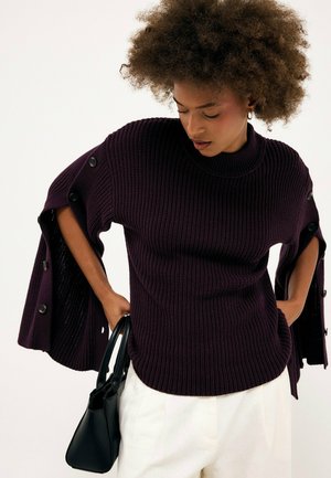 Femme portant un pull côtelé violet foncé avec des manches ouvertes boutonnées, un pantalon blanc, tenant un sac à main noir, regardant vers le bas.