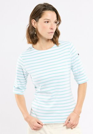 Femme aux cheveux bruns mi-longs portant des boucles d'oreilles dorées et une chemise blanche à rayures horizontales bleu clair, regardant sur le côté.