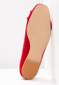 Rode suède platte schoen met een gladde, ronde neus en een platte, ribbelige rubberen zool. De onderkant is beige met "MADE IN SPAIN" ingeschreven.