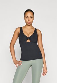 Fekete, testhezálló tank top elöl csavart dísz�ítéssel és kulcslyuk kivágással; világoszöld leggingssel párosítva, látható varrás részletekkel.