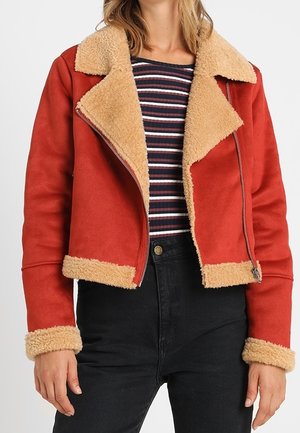 Frau trägt eine rote Lammfelljacke mit beige-farbener Fellfütterung, ein gestreiftes Hemd und schwarze Jeans mit hoher Taille, sie steht vor einem schlichten Hintergrund.