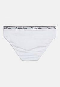 Λευκό σλιπ μπικίνι Calvin Klein με λογότυπο στη μέση σε μαύρο κείμενο πάνω σε γκρι φόντο, επίπεδη διάταξη πάνω σε λευκή επιφάνεια.