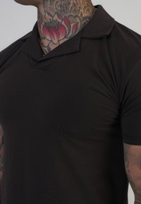 Čierne polo tričko s jemnou, hladkou textúrou, so klasickým golierom a krátkymi rukávmi, tesne priliehajúce k trupu.