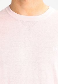 Nahaufnahme einer Person, die einen hellrosa Rundhalsausschnitt-Pullover trägt, mit einem kleinen strukturierten Dreieckdetail unter dem Kragen.