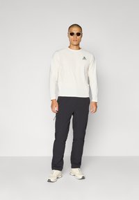Crèmekleurig overhemd met lange mouwen en een driehoekig logo, gecombineerd met een donkere broek met zijritsen en beige sneakers. Man draagt een zonnebril.