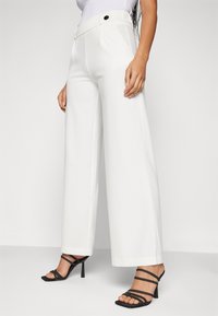 Pantaloni bianchi a gamba larga con una texture liscia, vita alta e pieghe frontali. Abbinati a sandali neri con tacco alto e cinturini.
