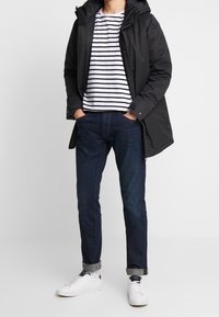 Svart huva parkas, vit och marinrandig t-shirt, mörka denimjeans, vita sneakers. Jackan har tryckknappar och fickor.