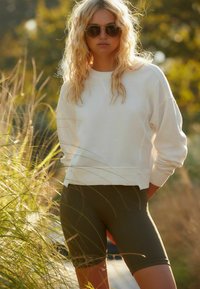 Witte sweatshirt met geribbelde textuur en een iets verkorte zoom, gecombineerd met donkergroene, strakke fietsbroekjes. Model draagt ronde zonnebril.