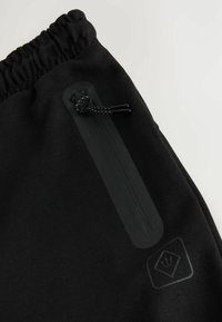 Detail černých kalhot s elastickým pasem, svislým zipovým kapsou a malým vyraženým logem u kapsy.