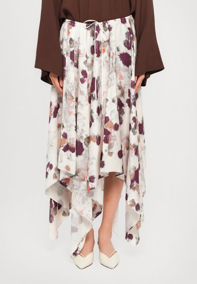 Jupe blanche fluide asymétrique avec des motifs floraux violets et oranges, portée avec un haut marron à manches longues et des chaussures blanches à bout pointu.