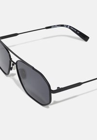 Melni saulesbrilles ar sešstūru rāmi, tumši pelēkiem stikliem un šaurām melnām metāla kājām. Rāmī ir teksturēti akcenti.