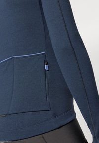Tums tumši zilā krāsa garām piedurknēm sporta krekls ar rāvējslēdzēja sānu kabatu, teksturētu audumu, kontrastējošu gaiši zilu šuvēju un pieguļošu dizainu.