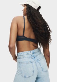 Frau mit langen lockigen Haaren, die eine weiße Mütze, einen dunklen BH und hoch taillierte hellblaue Jeans aus Denim trägt, steht mit dem Rücken zur Kamera.