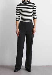 Pantalon noir à jambes larges avec une texture lisse, associé à un col roulé rayé noir et blanc. Le modèle est debout, les mains sur les hanches.