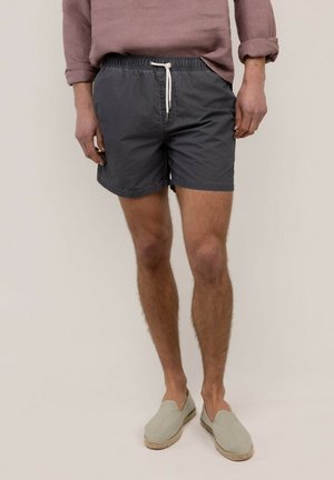 Hombre vestido con pantalones cortos ajustables de color gris oscuro, zapatos beige claro sin cordones y una camisa de manga larga malva con las mangas remangadas.