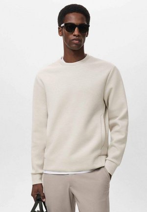 Sudoes de color crema con mangas largas, cuello redondo, bolsillos laterales y una textura suave, combinados con gafas de sol negras y pantalones claros.