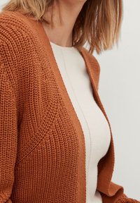 Strickjacke in rostorange mit offenem Vorderteil und strukturiertem Muster. Getragen über einem hellbeigen Oberteil mit subtilen Nahtdetails.