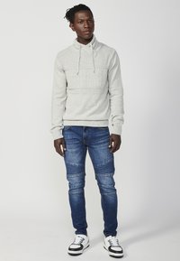 Helle graue Kapuzenjacke mit Textur auf der Vorderseite, Kordeln und gerippten Bündchen, kombiniert mit blauen Skinny-Jeans mit Panelelementen und weißen Sneakers.