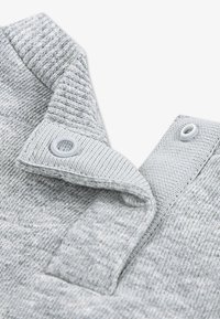 Γκρι πλεκτό ύφασμα με υφασμένο μοτίβο, διαθέτοντας γιακά και δύο κλ ιπ στερέωσης κοντά στον λαιμό.