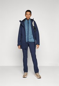 Marine vanntett jakke med glidelås, fleecefôr og hette, kombinert med en blå fleecegenser, marineblå bukser og beige joggesko.