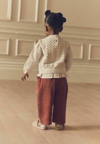Barn iført en strikket cremet vest med et herringbone-mønster, parret med rustfarvede fløjlsbukser og beige slip-on sko.