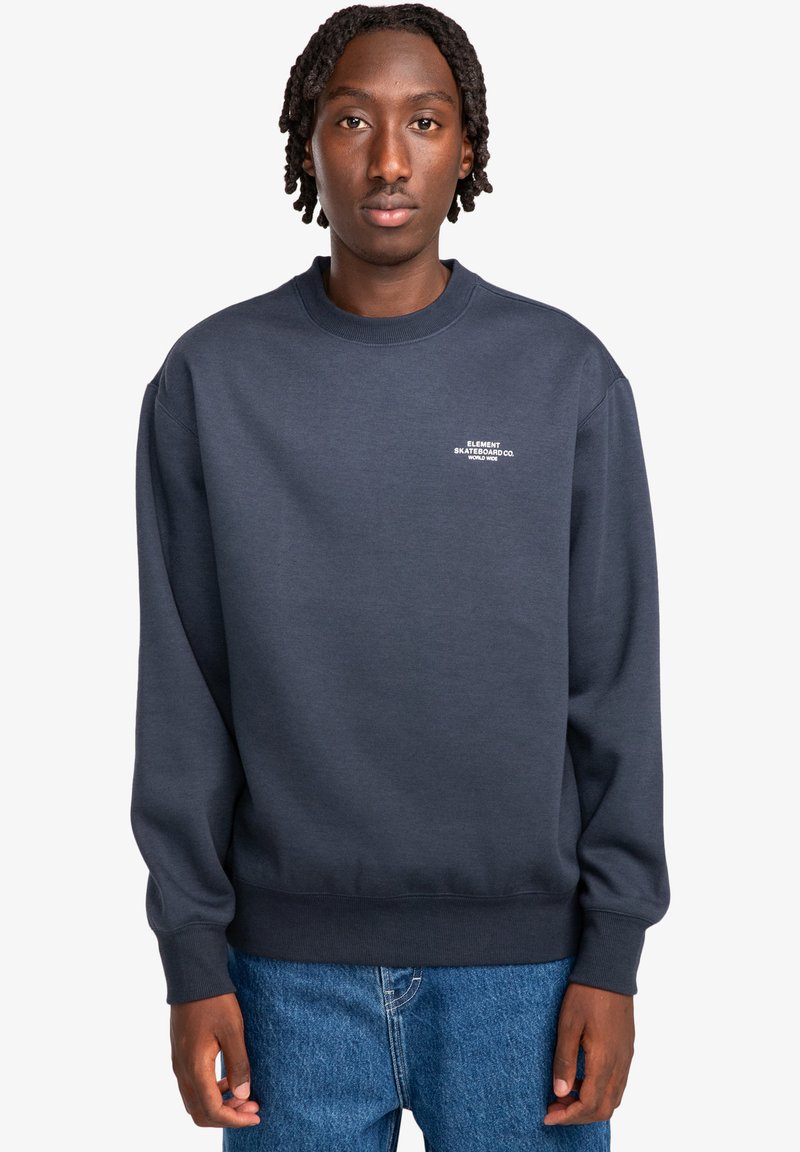 Felpa crewneck blu in morbido tessuto con un piccolo logo bianco sul petto e polsini e orlo a costine. Indossata con jeans blu.