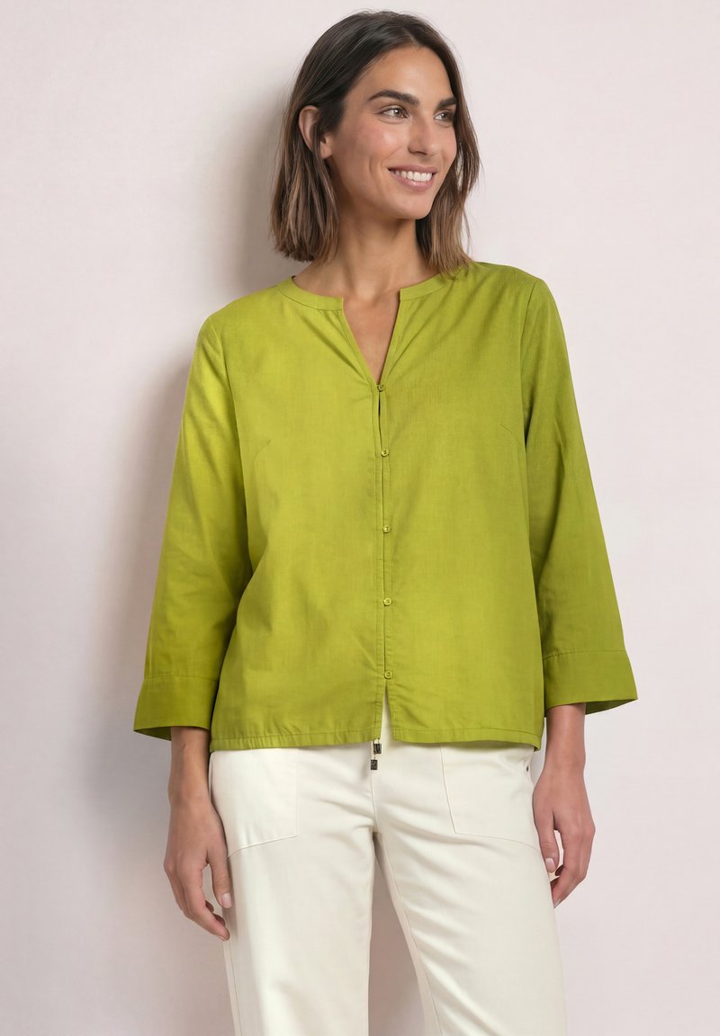 Femme souriante, portant un chemisier boutonné à manches longues vert citron vert et un pantalon blanc, debout devant un fond clair uni.