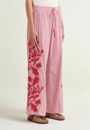 Pantaloni a gamba larga a righe verticali rosse e bianche con ricamo floreale rosso sul lato, indossati con sandali marroni e una maglietta bianca.