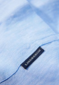 Tela de lino azul claro con textura visible, que presenta una etiqueta azul oscura con "LINDBERGH" en texto blanco cosido en el borde.