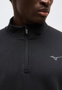 Κοντινό πλάνο ενός ατόμου που φοράει μαύρο τοπ με φερμουάρ και ψηλό γιακά, με λογότυπο στο αριστερό στήθος.