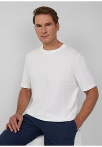 Weißes, kurzärmliges Baumwoll-T-Shirt mit Rundhalsausschnitt und kleinem Logo in heller Farbe, kombiniert mit navyfarbener Hose. Glatte Textur.