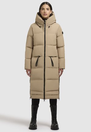 Beige puffer-takki, jossa on korkea kaulus ja suuri huppu. Siinä on vaakasuorat saumat, mustat vetoketjut ja sivutaskut. Yhdistetty mustien saappaitten kanssa.