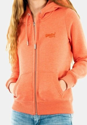 Mujer con sudadera naranja con cremallera, bolsillos delanteros y cordones, combinada con vaqueros azul claro, mostrada desde los hombros hasta las caderas.