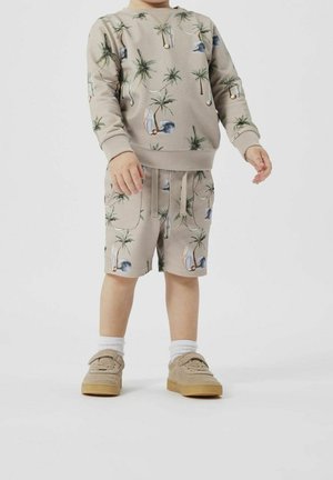 Tout-petit portant un short beige et un sweat-shirt avec un motif de palmier et de voilier, des chaussettes blanches et des baskets beige, debout sur un fond blanc.