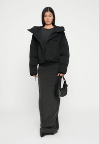 Giubbotto imbottito nero con design accorciato, abbinato a un lungo vestito grigio scuro. La modella tiene una piccola borsa nera texturizzata.