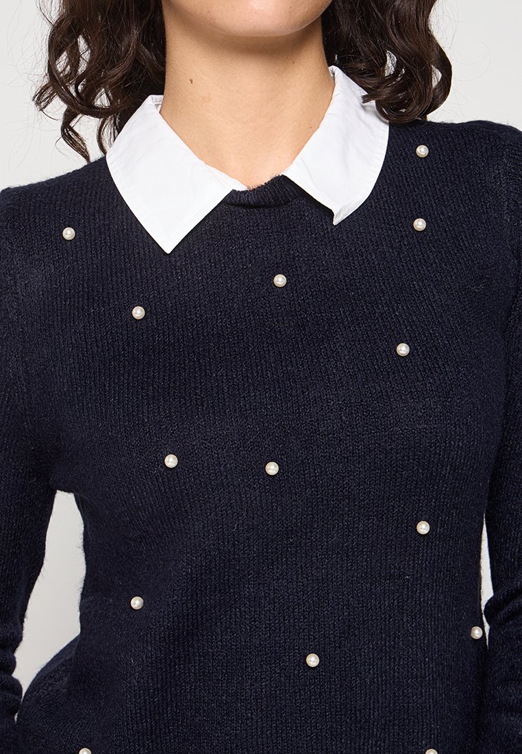 Pull en tricot bleu marine avec un col rond, une chemise blanche en dessous et des perles décoratives uniformément réparties sur le devant.