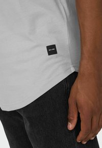 Light gray cotton t-shirt with short sleeves, featuring a stitched black label at the hem. Paired with dark denim pants.