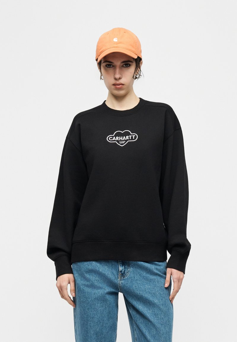 Persona con indosso un cappello arancione, una felpa nera Carhartt WIP e jeans blu, su uno sfondo chiaro e uniforme.