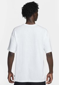 Man med mörk hy och korta dreadlocks som bär en enkel vit t-shirt med kort ärm, sedd bakifrån mot en vit bakgrund.