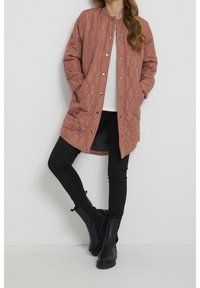 Femme portant un long manteau matelassé rose poussiéreux, un haut blanc, des leggings noirs et des bottines noires, se tenant avec une jambe croisée sur l'autre.